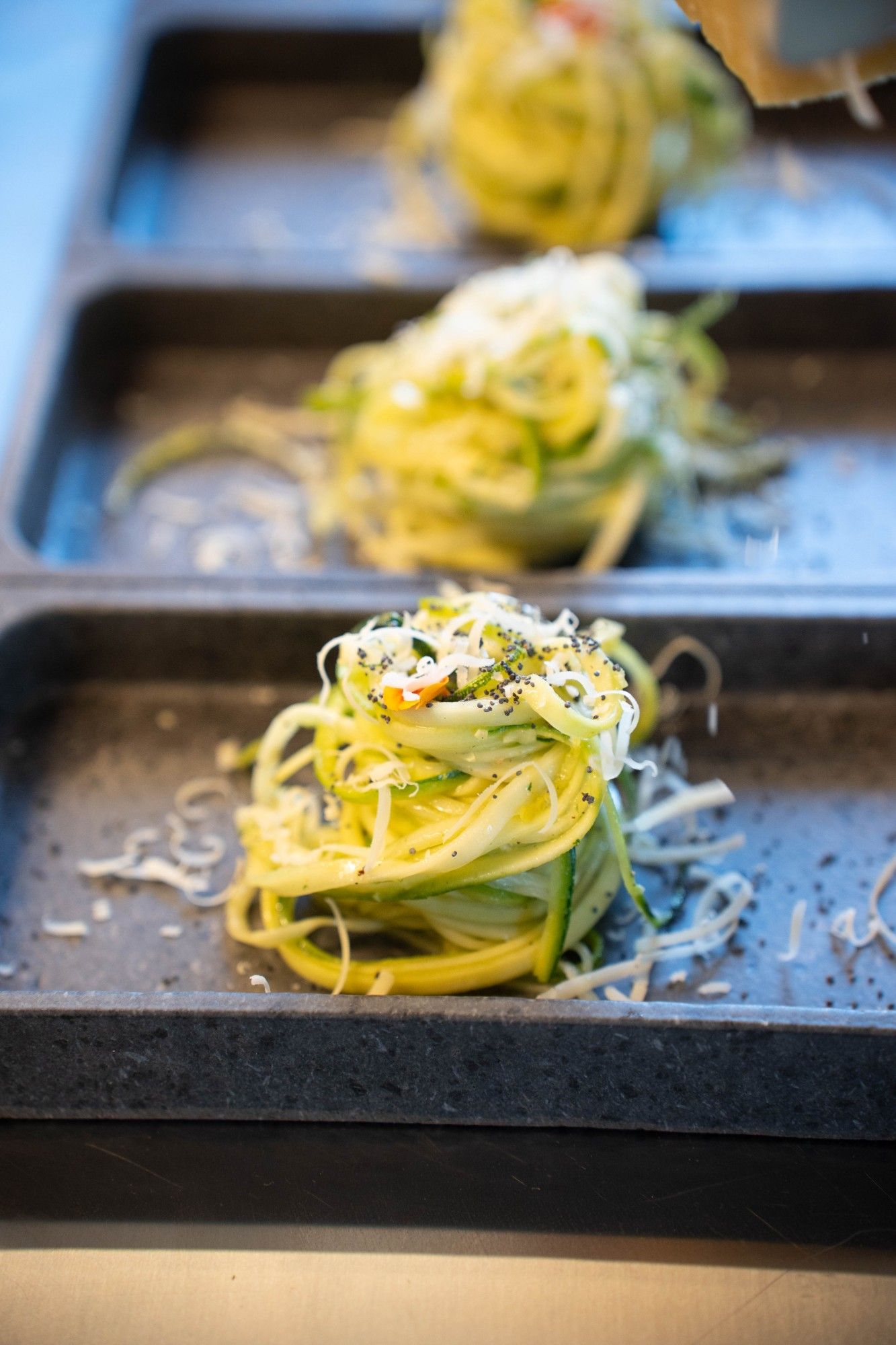 Nidi di spaghetti di zucchine con formaggio grattugiato e semi di papavero, impiattati su vassoi di pietra scura
